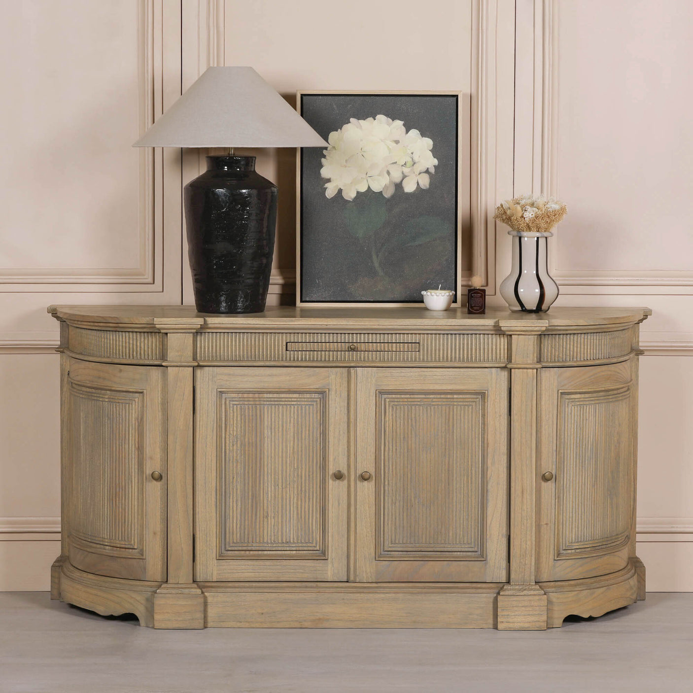 Rustic wooden buffet sideboard with fluted detailing, styled with a lamp, vase, and framed artwork against a neutral wall.