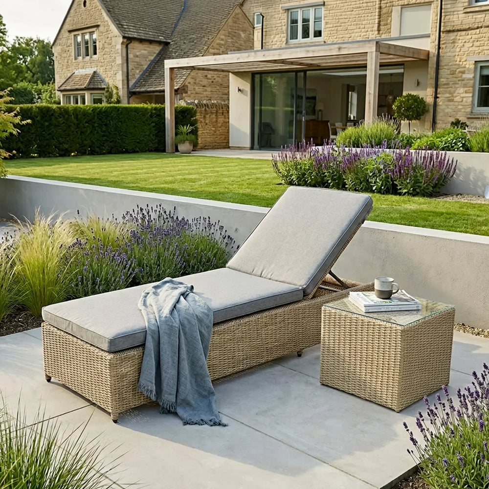Rattan sun lounger with grey cushions and throw beside matching side table in modern garden