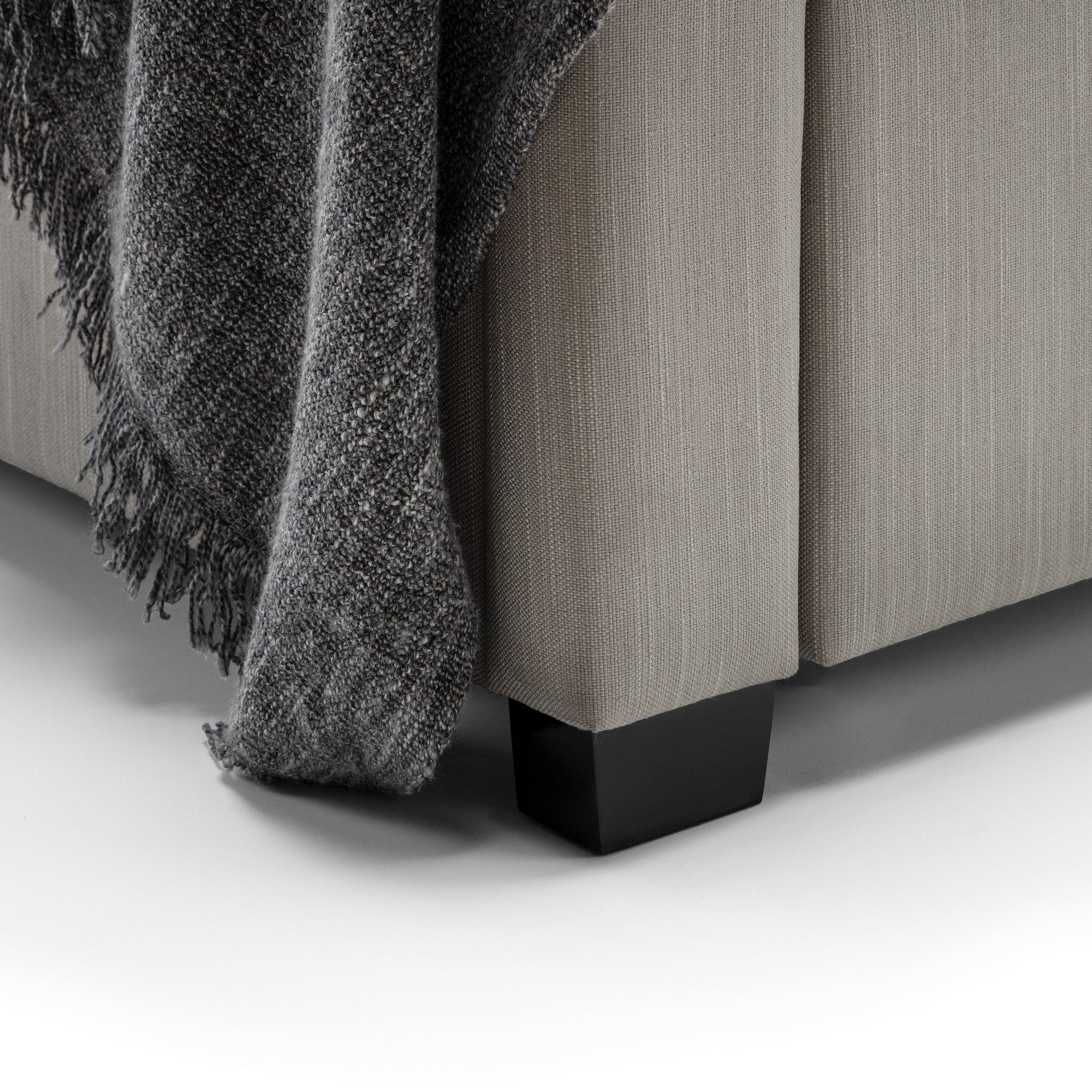 Foot detail of upholstered bed frame with dark wood leg and grey throw blanket