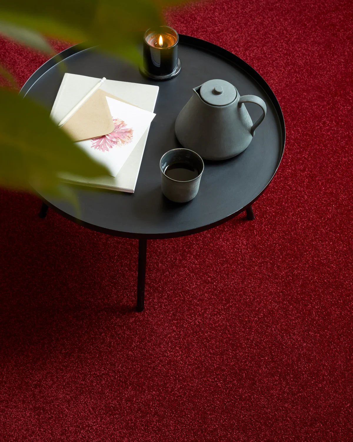 Round black coffee table on plush red carpet with teapot, cup, notebook and candle