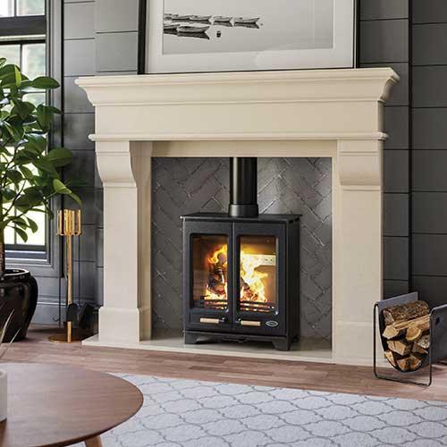 Modern wood burning stove with glass doors in stone fireplace, logs stacked nearby