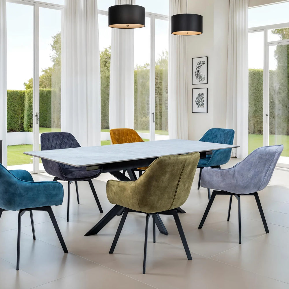 A modern dining room scene with a rectangular white dining table surrounded by six upholstered chairs in blue, gray, yellow, and purple velvet, set in a bright room with large windows and outdoor views.