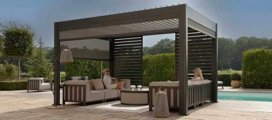 A large outdoor patio cabana with a built-in lounge set, shown in a landscaped backyard next to a swimming pool. Two people are seated inside the cabana under a dark gray slatted roof.