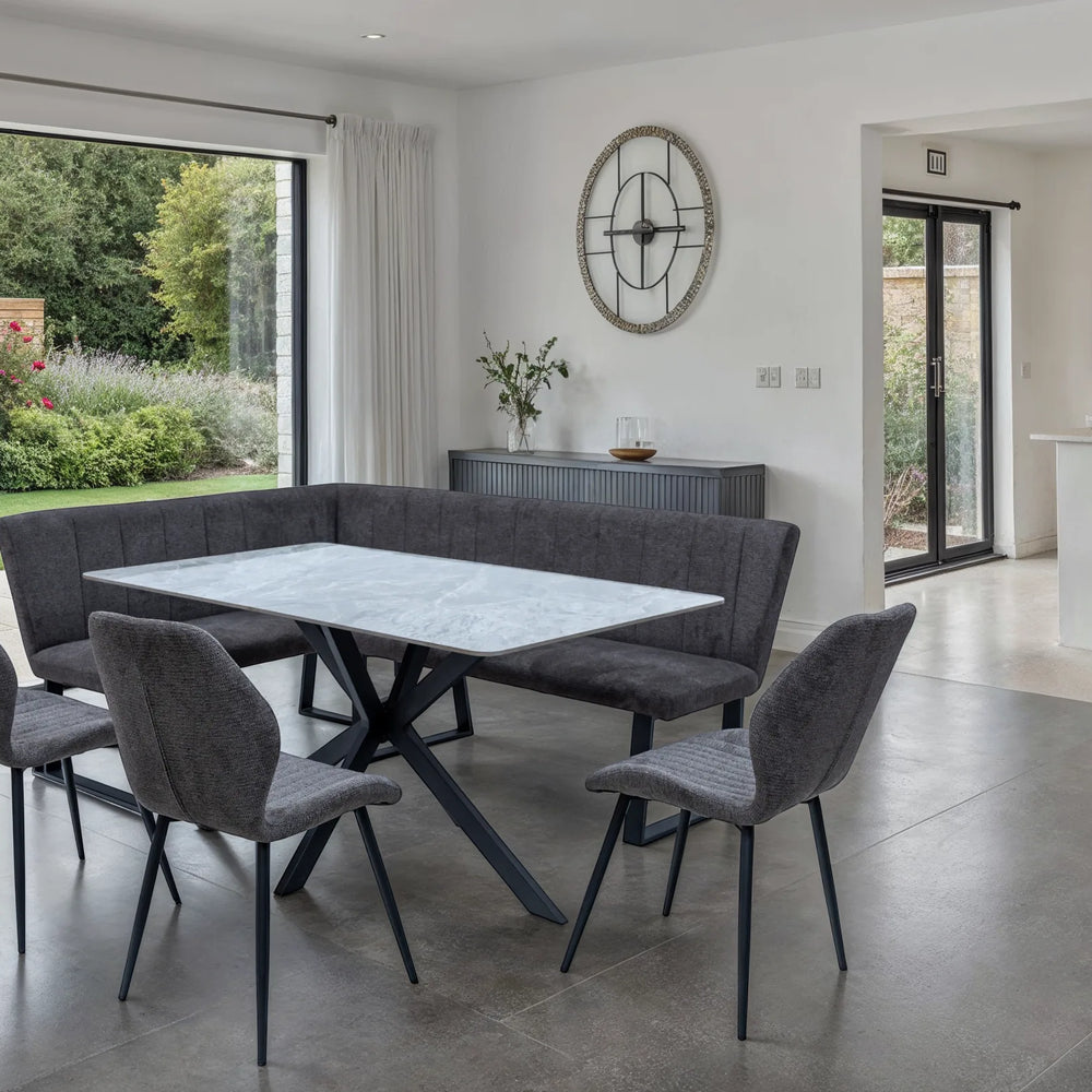 A modern dining set featuring a rectangular white marble-top table with black metal legs, surrounded by a dark gray upholstered corner banquette and four matching gray chairs, set in a bright contemporary dining room.