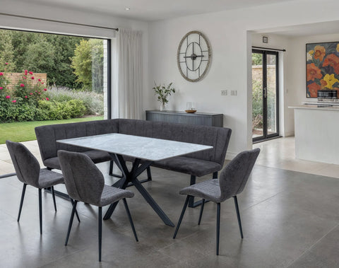 A modern dining set featuring a rectangular white marble-top table with black metal legs, surrounded by a dark gray upholstered corner banquette and four matching gray chairs, set in a bright contemporary dining room.
