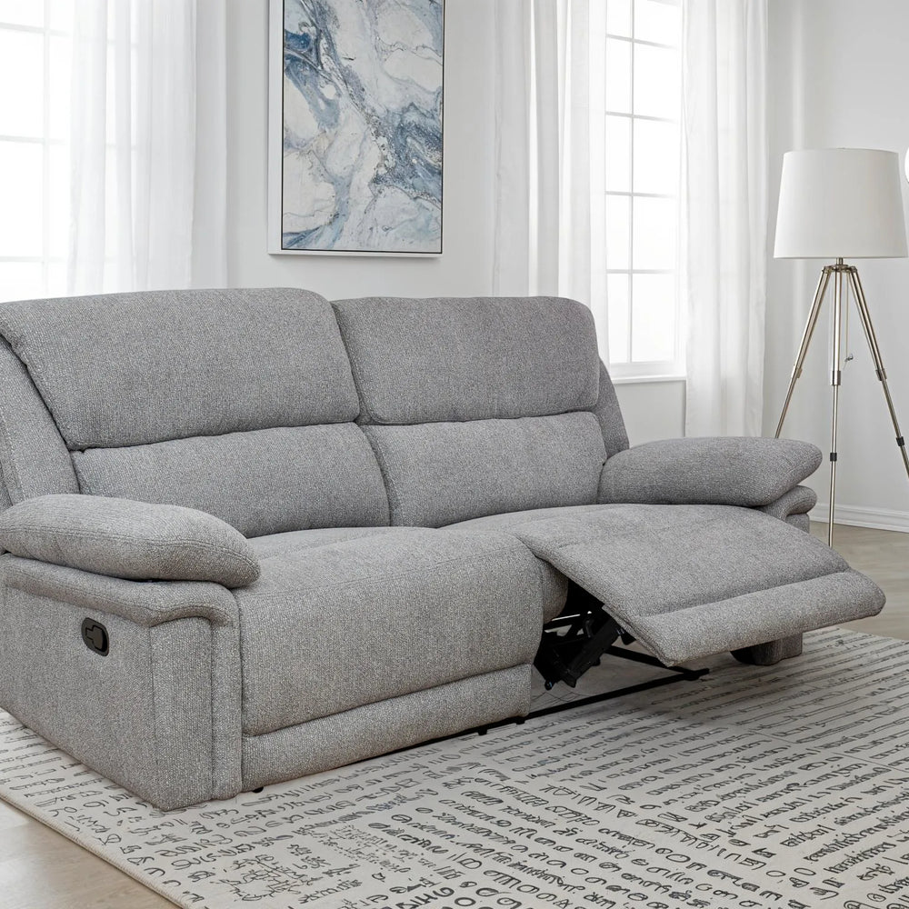 A light gray upholstered reclining loveseat sofa shown in a bright living room with windows, a floor lamp, wall art, and a patterned rug.