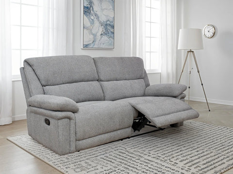 A light gray upholstered reclining loveseat sofa shown in a bright living room with windows, a floor lamp, wall art, and a patterned rug.