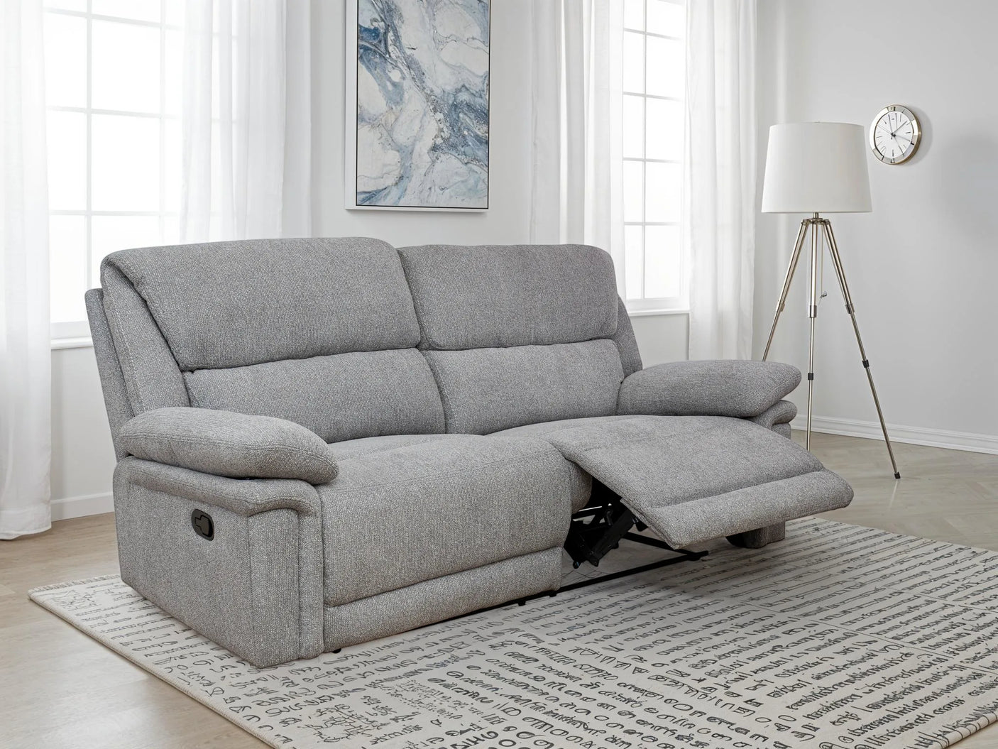 A light gray upholstered reclining loveseat sofa shown in a bright living room with windows, a floor lamp, wall art, and a patterned rug.
