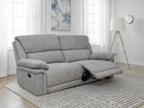 A light gray upholstered reclining loveseat sofa shown in a bright living room with windows, a floor lamp, wall art, and a patterned rug.