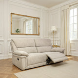 A light beige upholstered reclining loveseat sofa shown in a bright, white-paneled living room with a floor lamp, curtains, and a large gold-framed mirror.