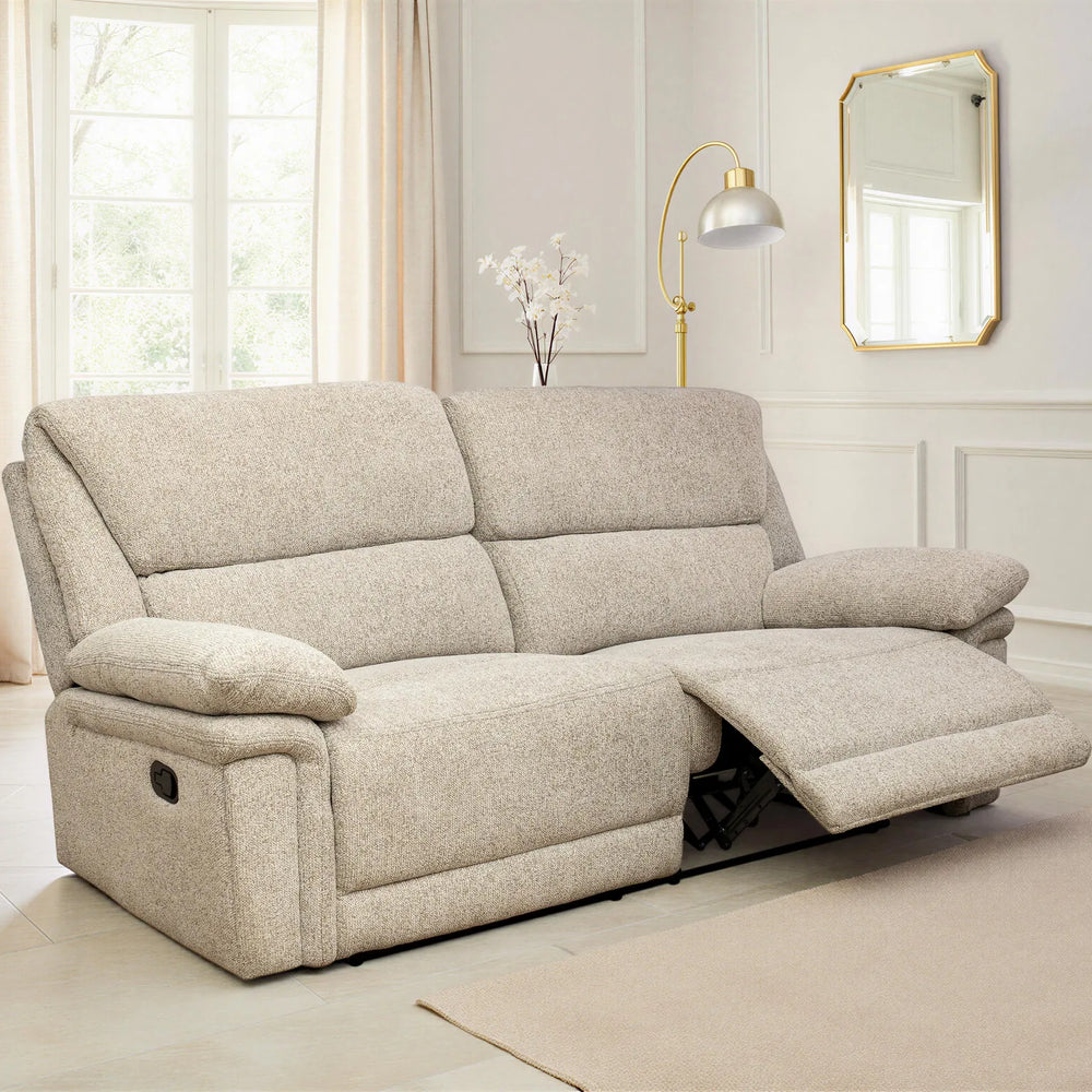 A light beige upholstered recliner sofa shown in a bright, modern living room with cream walls, curtains, a floor lamp, and a mirror. The sofa is shown in the reclined position with the footrest extended.