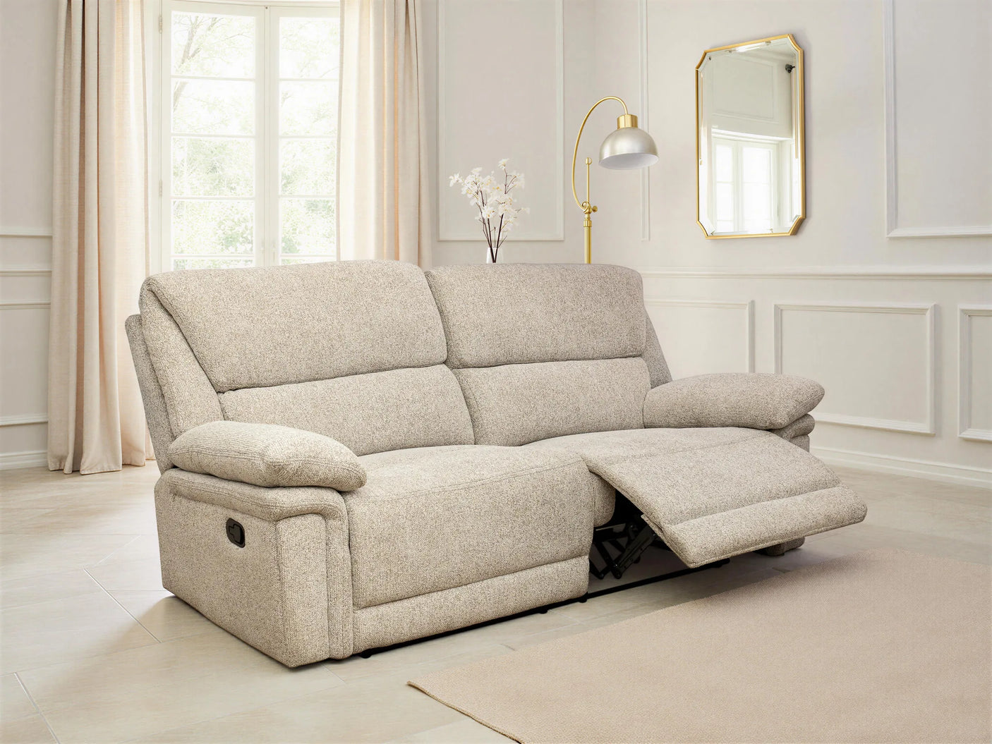 A light beige upholstered recliner sofa shown in a bright, modern living room with cream walls, curtains, a floor lamp, and a mirror. The sofa is shown in the reclined position with the footrest extended.