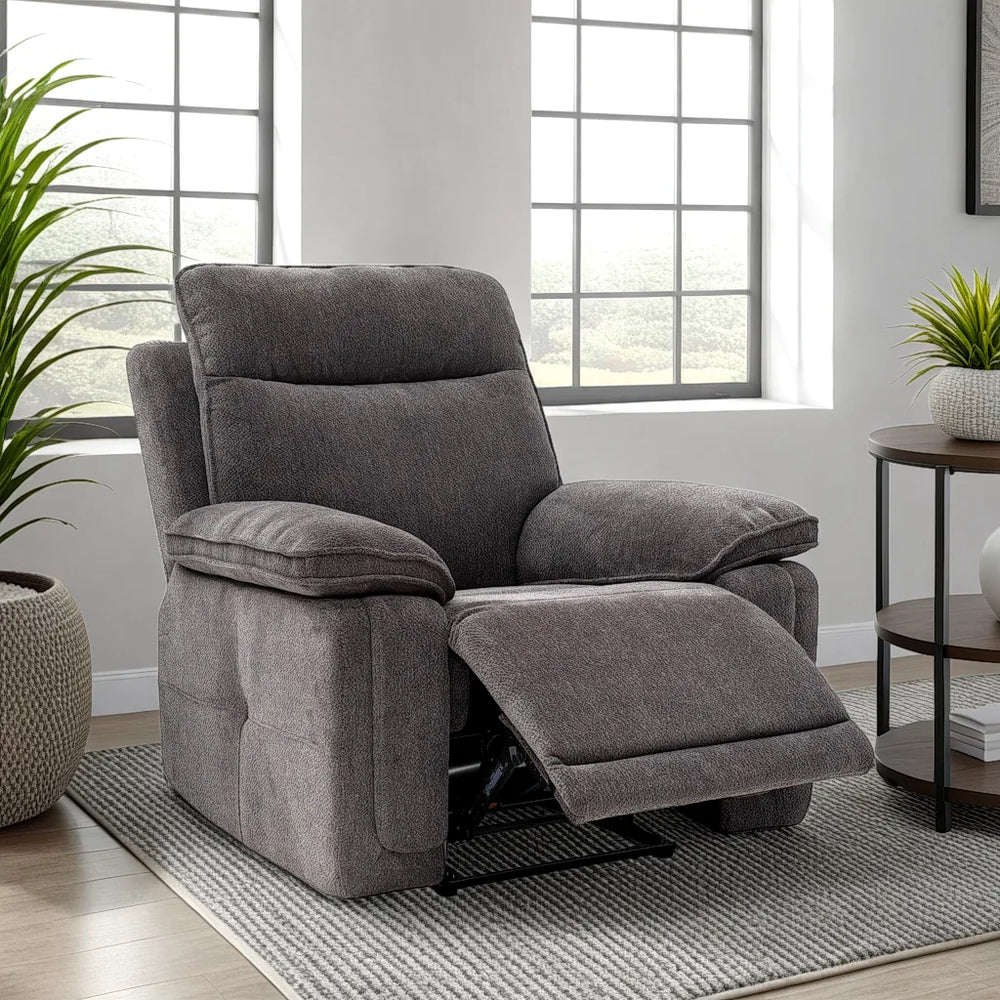 A gray fabric recliner chair with the footrest extended, positioned on a patterned rug in a bright living room setting with windows, plants, and a side table.