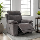 A gray fabric recliner chair with the footrest extended, positioned on a patterned rug in a bright living room setting with windows, plants, and a side table.