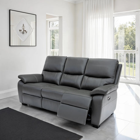 A three-seater gray leather reclining sofa with one seat cushion extended, positioned in a bright, modern living room with white walls, large windows, and a black rug in the foreground.