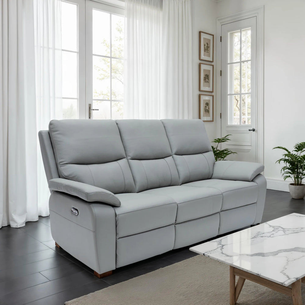 A light gray three-seat reclining sofa in a bright, modern living room with white curtains, dark wood floors, and a marble-top coffee table.