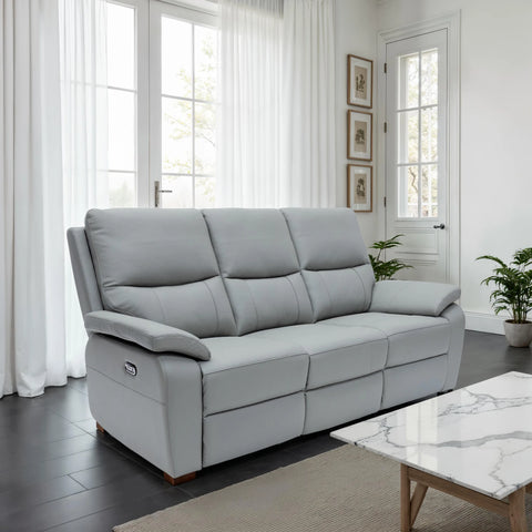 A light gray three-seat reclining sofa in a bright, modern living room with white curtains, dark wood floors, and a marble-top coffee table.