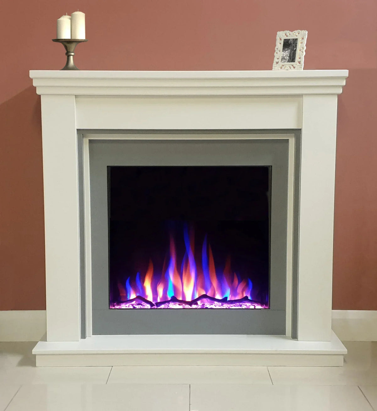 White electric fireplace with multicolored LED flames against pink wall, candles on mantel