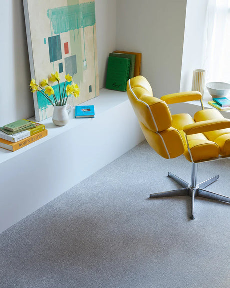 Yellow swivel chair with chrome base on light grey carpet in a bright room with books and art.