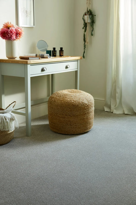Grey carpet in a bedroom with a woven pouf and light wooden vanity table
