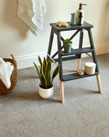 Dark grey step stool with natural wood legs, styled with plants and toiletries in a bathroom.