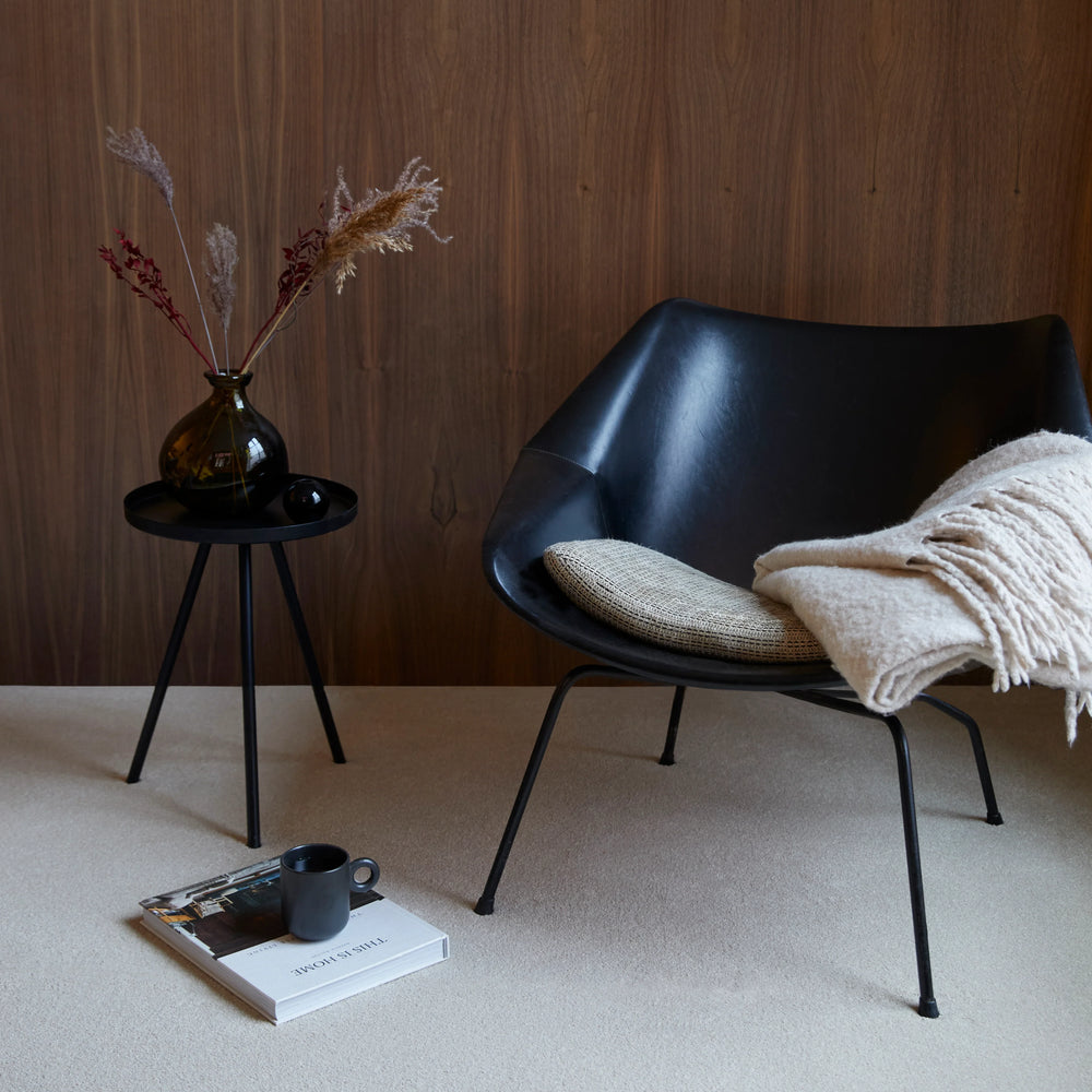 Black modern chair with a beige cushion, styled with a side table and vase on cream carpet