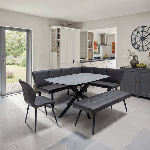 A modern dining set in a bright, contemporary dining room. The set includes a rectangular gray table with black crossed legs, a tufted corner banquette, a matching seat bench, and a gray upholstered chair with black legs.