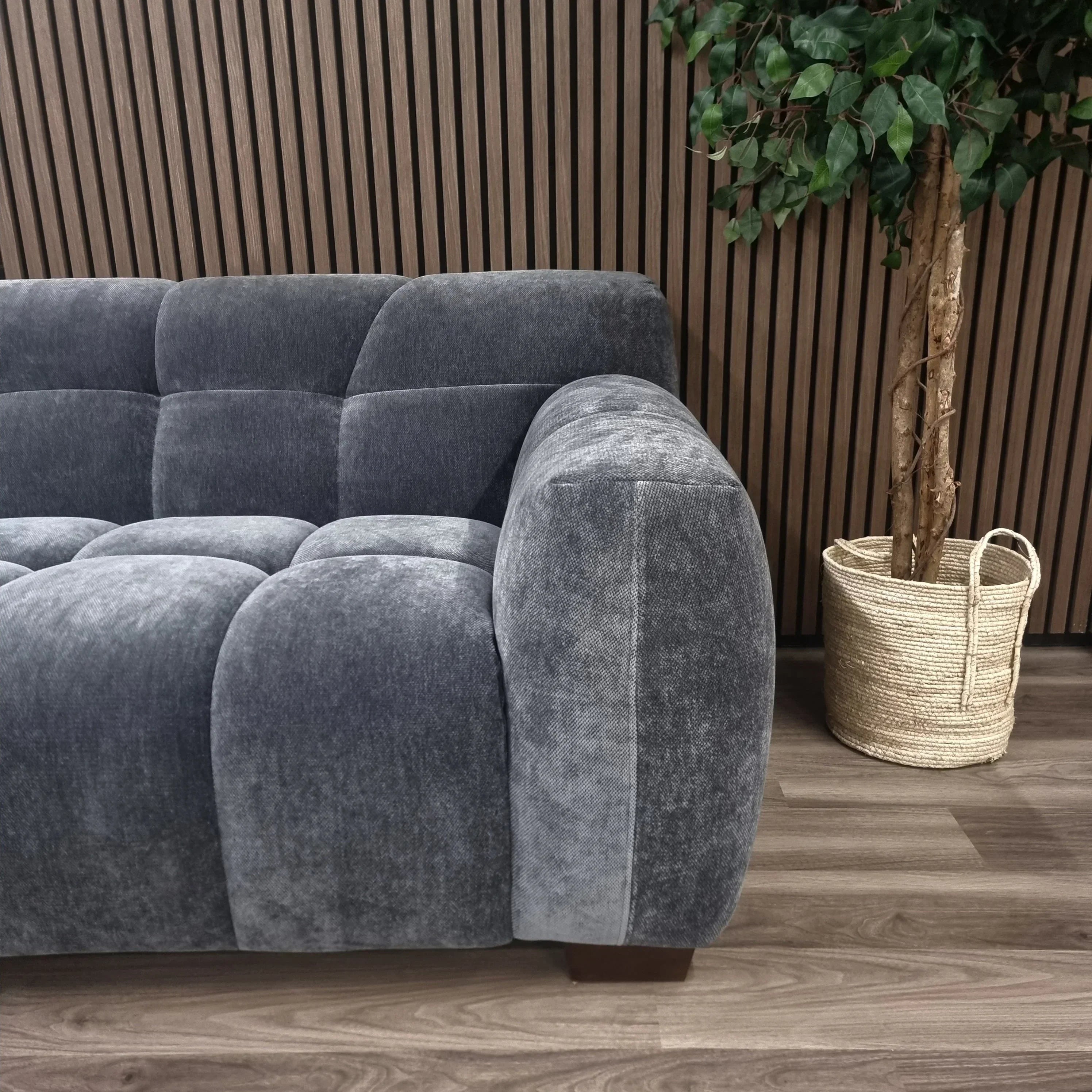 Grey fabric corner sofa with tufted cushions beside potted plant, wood panel backdrop