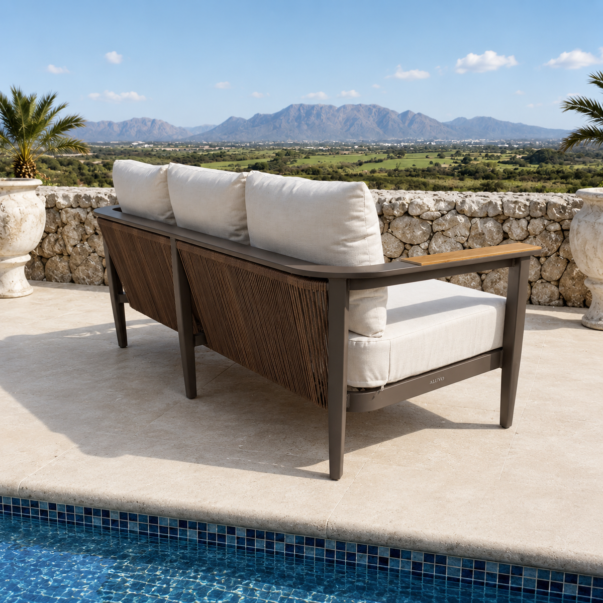 Beige cushioned sofa with dark brown metal frame beside a blue tiled pool and stone wall.