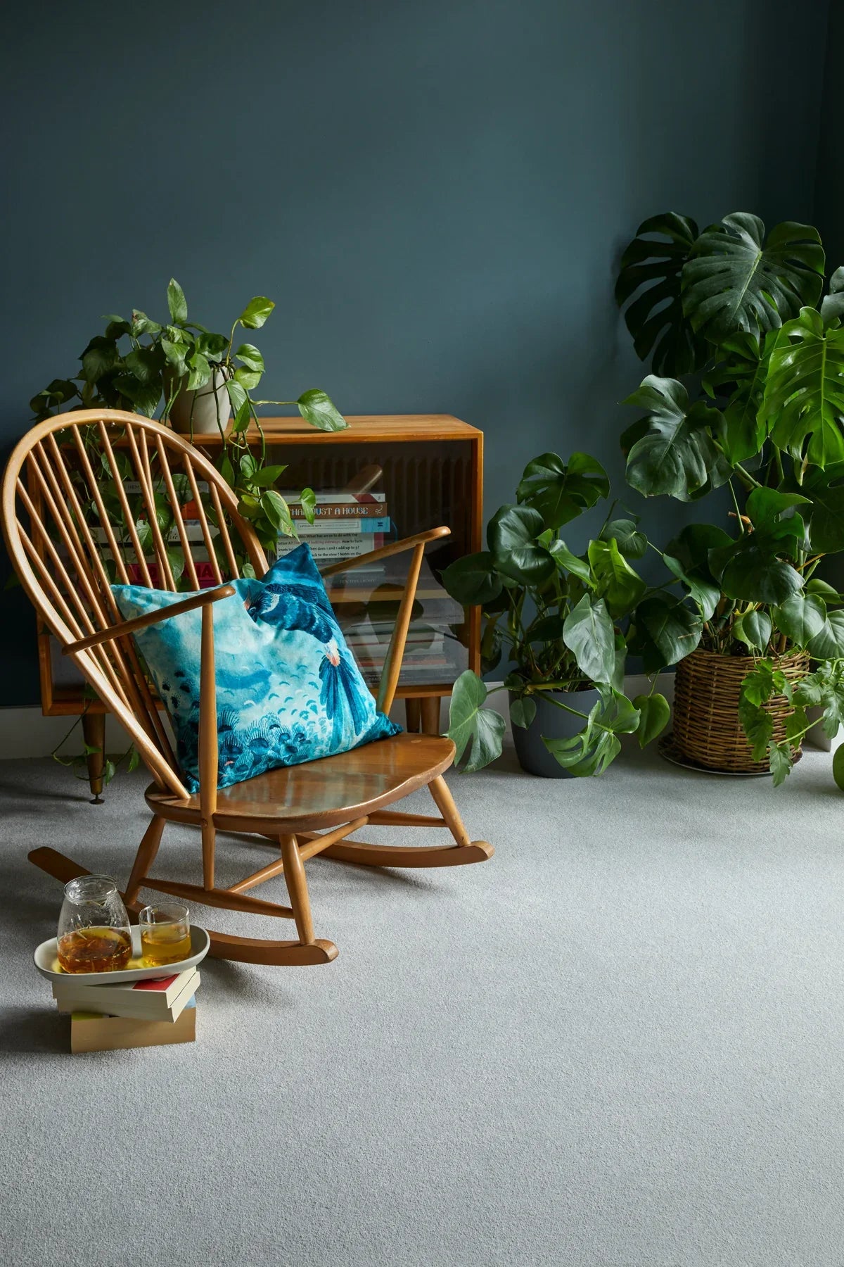 Wooden rocking chair with blue patterned cushion in a room with plants and books.