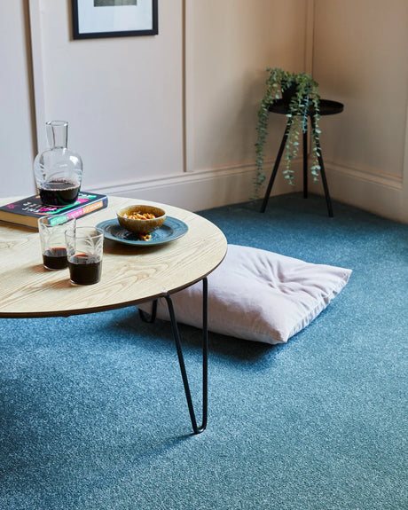 Round light wood table with black metal legs on blue carpet in cozy living room scene