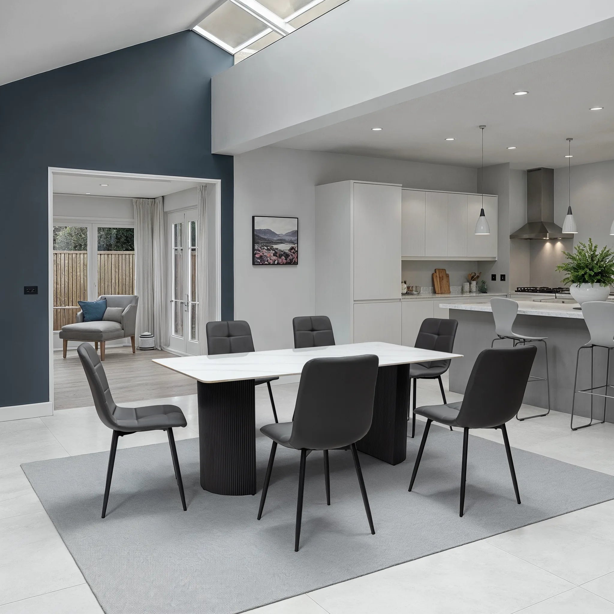 A modern dining room setup featuring a rectangular white-top dining table with six dark gray upholstered chairs, placed on a light gray rug in an open-plan kitchen and living space.