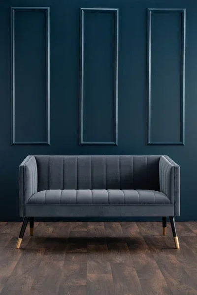 Dark grey velvet sofa with gold-tipped legs against blue paneled wall and wood floor