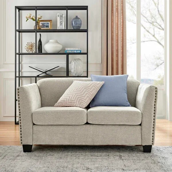 Beige fabric sofa with studded arms and two cushions in living room with black shelving unit