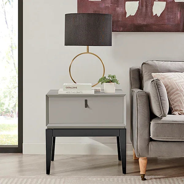 Grey bedside table with drawer beside sofa, topped with lamp, books, and small plant