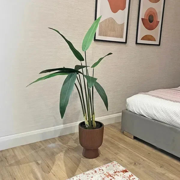 Tall artificial plant in ribbed brown planter beside bed and wall art in modern bedroom