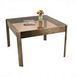 Square glass dining table with brushed wood frame, topped with a small vase and magazines