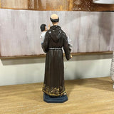 Brown religious statue with gold trim on wooden table, figure holding child and lily flower
