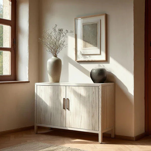 Light wood sideboard with two doors, decorative vases, and framed art in a sunlit room corner