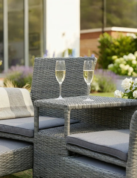 Grey rattan garden sofa set with glass table, two champagne flutes, and seat cushions