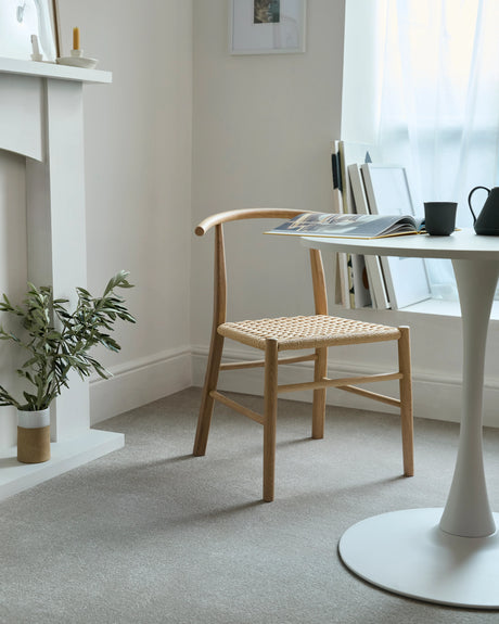Natural wood chair with woven seat next to a white round table in a bright, airy room.