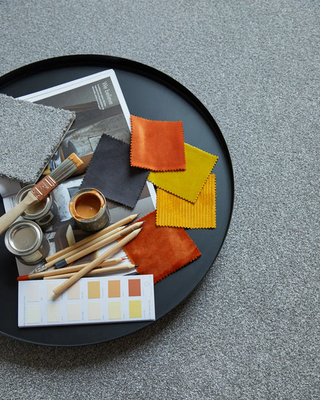 Round black tray with vibrant fabric swatches, paintbrushes, and paint samples on grey carpet