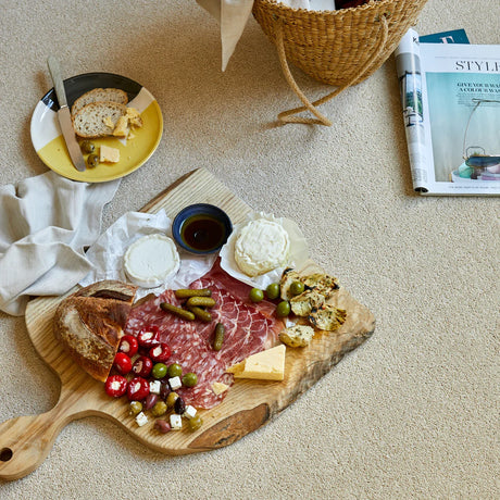 Wooden serving board with assorted cheeses, cured meats, and olives on a beige carpet.