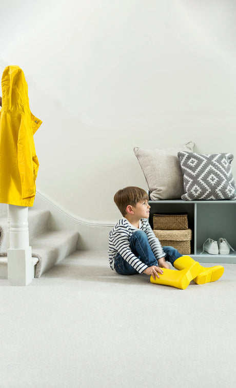 Child in striped shirt sitting on light grey carpet with yellow boots, next to storage with