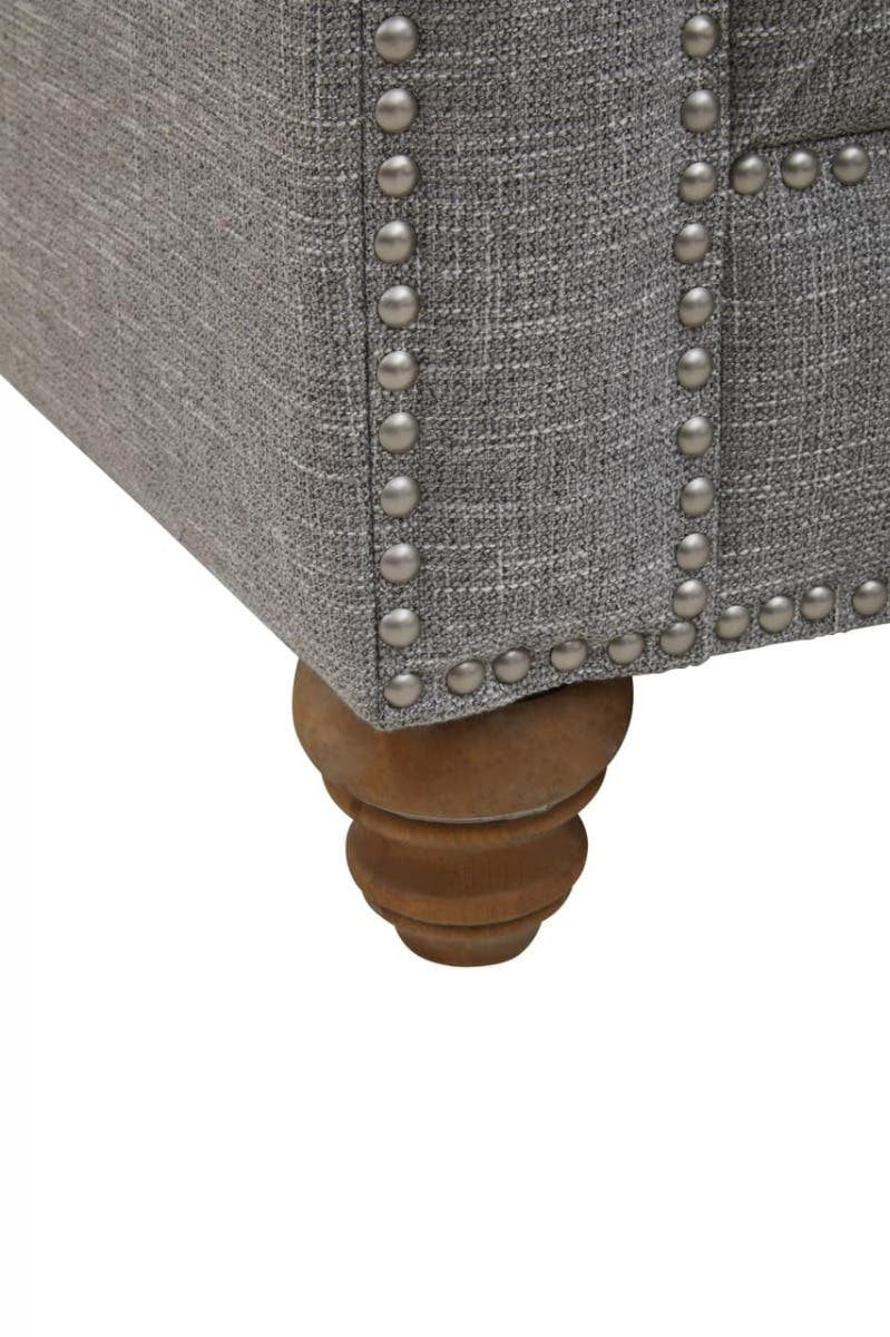 Close-up view of the wooden leg of a grey linen sofa, featuring a rounded design and decorative nailhead trim along the edge of the upholstery