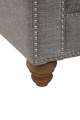 Close-up view of the wooden leg of a grey linen sofa, featuring a rounded design and decorative nailhead trim along the edge of the upholstery